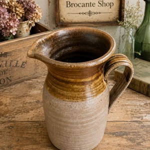 May include: A ceramic pitcher featuring a two-tone glaze with a dark brown upper section and a textured, light beige lower body. The vessel has a wide, rounded rim with a prominent pouring spout and a sturdy, curved handle on the right side.