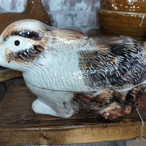 May include: A ceramic rabbit-shaped container with a lid. The container is primarily white with brown and black markings, and a glossy finish. The rabbit is posed in a seated position, with its ears laid back. The container is sitting on a wooden surface.