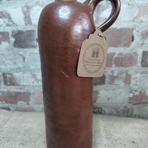 Op de afbeelding: Een bruine steengoed kruik met een handvat en een kleine tuit. De kruik heeft een getextureerd oppervlak en een label met de tekst "The Little Brocante Shop". Het is een decoratief item in vintage stijl.