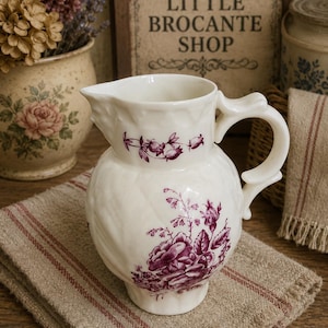 May include: A cream-colored ceramic pitcher featuring a textured, embossed surface and a deep purple floral transferware pattern. The pitcher has a curved handle, a wide spout, and a rounded body decorated with detailed roses and foliage.
