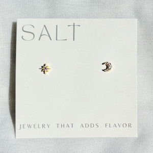 May include: A pair of gold-tone stud earrings on a white display card. One earring is shaped like a star with a central clear stone, and the other is a crescent moon with small clear stones. The card features the text SALT and JEWELRY THAT ADDS FLAVOR.