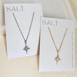 May include: Two necklaces on white cards. One is silver-toned, the other gold-toned. Each features a delicate chain and a starburst pendant with a central clear stone. The cards are labeled "SALT" and "WITH LOVE FROM THE CALIFORNIA COAST."