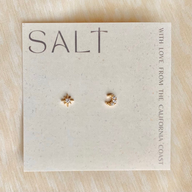 Dainty gold star and moon stud earrings with sparkling cubic zirconia stones, displayed on a neutral-toned 'SALT' jewelry card. Minimalist celestial earrings perfect for everyday wear or gifting.