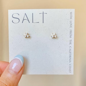 May include: A pair of gold-toned stud earrings, each featuring a cluster of three clear, sparkling crystals. The earrings are displayed on a white card with the word "SALT" printed in gray and the text "WITH LOVE FROM THE CALIFORNIA COAST".