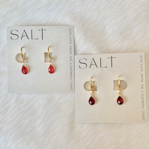 May include: Two pairs of gold-tone earrings with teardrop-shaped, red gemstones. The earrings are displayed on white cards with the word "SALT" printed in black. The cards also include the text "WITH LOVE FROM THE CALIFORNIA COAST."