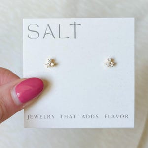 May include: A pair of gold-toned stud earrings, each featuring three small clear stones arranged in a triangular cluster. The earrings are mounted on a white display card with the text SALT and JEWELRY THAT ADDS FLAVOR printed in gray.