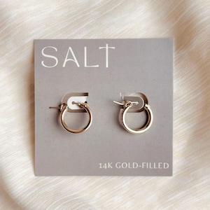 May include: A pair of small gold hoop earrings attached to a gray rectangular display card. The card features the word SALT printed in white at the top and the text 14K GOLD-FILLED printed in white at the bottom center of the display card.
