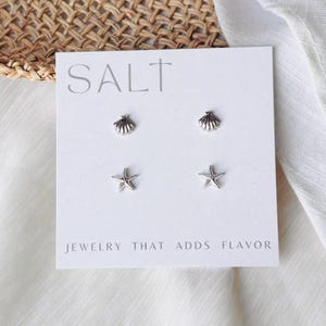 May include: A white card with the word "SALT" and two pairs of silver earrings: seashell and starfish shapes. The text "JEWELRY THAT ADDS FLAVOR" is printed below the earrings. The background includes a woven texture and white fabric.