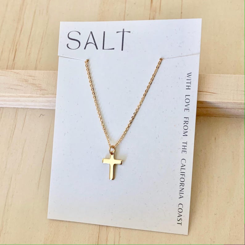 May include: A gold-colored cross pendant necklace on a white card with the word "SALT" and the text "WITH LOVE FROM THE CALIFORNIA COAST". The necklace is displayed on a light wooden surface.
