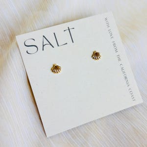 May include: Gold-colored seashell stud earrings on a white card with the word "SALT" and the text "WITH LOVE FROM THE CALIFORNIA COAST". The earrings are small and detailed, with a textured surface.