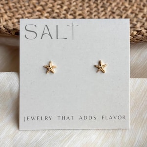 May include: A pair of gold-toned starfish stud earrings displayed on a rectangular white card. The card features the text SALT at the top and the phrase JEWELRY THAT ADDS FLAVOR at the bottom. The earrings are centered on the card.
