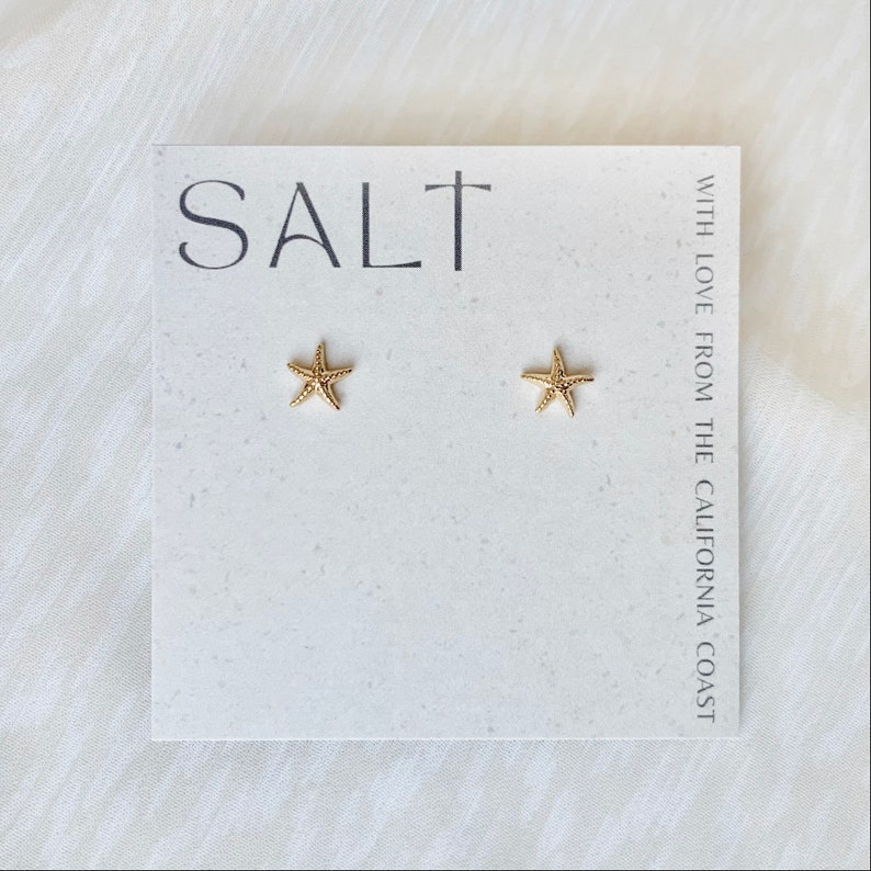 A photo featuring a pair of dainty, gold-plated starfish stud earrings displayed on a neutral "SALT" branded earring card. Lightweight sea star stud earrings are great for everyday wear or as a gift for her.