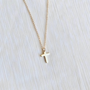 May include: A gold-colored necklace with a cross pendant. The cross is a simple, classic design, and the chain is delicate. The necklace is displayed against a white, textured background.
