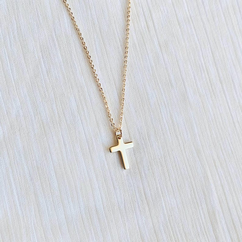May include: A gold-colored necklace with a cross pendant. The cross is a simple, classic design, and the chain is delicate. The necklace is displayed against a white, textured background.