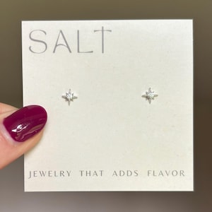 May include: A pair of silver-toned stud earrings with a starburst design, each featuring a central clear gemstone. The earrings are displayed on a white card with the word "SALT" at the top and the phrase "JEWELRY THAT ADDS FLAVOR" at the bottom.