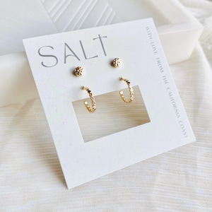 May include: A set of gold-colored earrings on a white card. The card has the word "SALT" printed in gray. The set includes two pairs of earrings: small stud earrings and hoop earrings. The hoop earrings have a dotted design. The card also has the text "WITH LOVE FROM THE CALIFORNIA COAST".