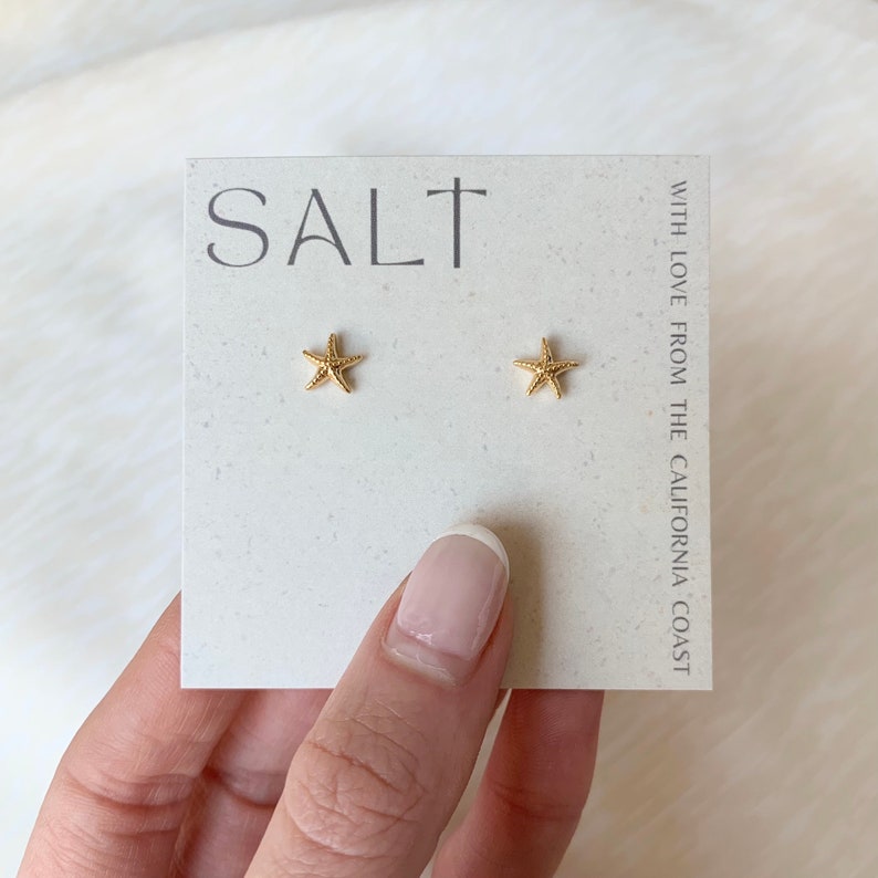 A photo featuring a pair of dainty, gold-plated starfish stud earrings displayed on a neutral "SALT" branded earring card being held by woman's hand. Cute, lightweight sea star stud earrings are great for everyday coastal jewelry or as a gift for a beach loving friend.