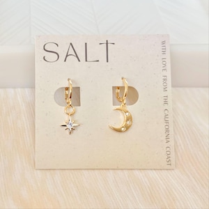 May include: Gold-colored hoop earrings. One earring features a star pendant with a central clear stone, and the other has a crescent moon with three clear stones. The earrings are displayed on a cream-colored card with the word "SALT" and the text "WITH LOVE FROM THE CALIFORNIA COAST".
