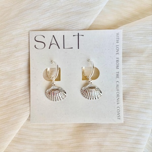 May include: A pair of silver hoop earrings with seashell charms. The earrings are displayed on a white card with the word "SALT" and the text "WITH LOVE FROM THE CALIFORNIA COAST". The background is a cream-colored fabric.