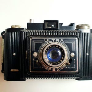 May include: A vintage black Fex Ultra camera with a silver lens and dials. The camera has the word "ULTRA" above the lens and "FEX" below. The camera has the word "HIMALAYA" on the side.