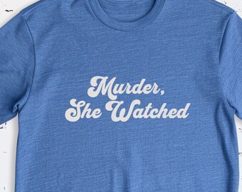 Op de afbeelding: Een blauw t-shirt met de tekst "Murder, She Watched" in witte retro-lettering.