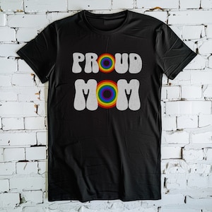 Pode incluir: Camiseta preta com o texto "PROUD MOM" em branco com um círculo arco-íris no meio da palavra "MOM".