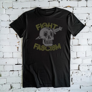 Könnte beinhalten: Schwarzes T-Shirt mit einem Grafikdruck eines Schädels mit einem Dolch, der durch ihn hindurchgeht. Der Text "FIGHT FASCISM" ist in Gelb um den Schädel herum geschrieben.