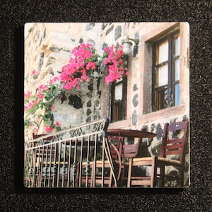 May include: A square image of a scenic outdoor cafe. The scene features a stone wall adorned with vibrant pink flowers. Wooden tables and chairs are set on a small balcony with a metal railing, creating a charming, inviting atmosphere.