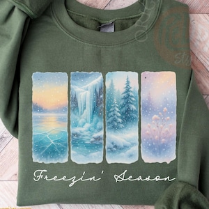 May include: Military green sweatshirt featuring a winter scene design. The design includes four vertical panels depicting a snowy landscape with a frozen lake, waterfall, and evergreen trees. The words "Freezin' Season" are written in white script.