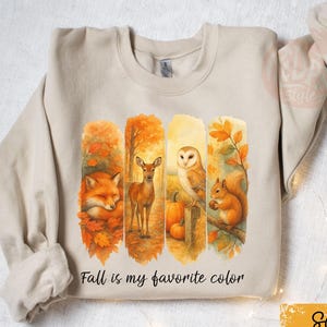 May include: A sand-coloured sweatshirt with a watercolour design of autumn animals: a fox, deer, owl, and squirrel. The design includes fall foliage and pumpkins. The text "Fall is my favourite colour" is printed below the animals.