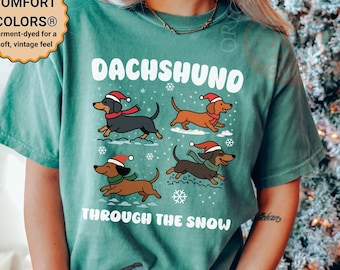 May include: Light green t-shirt with a Dachshund design. The shirt features four cartoon Dachshunds wearing Santa hats and scarves, with the text "DACHSHUND THROUGH THE SNOW". The shirt is garment-dyed for a soft, vintage feel.