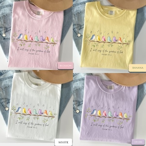 May include: Four short-sleeve t-shirts in blossom pink, banana yellow, white, and orchid purple. Each shirt features a graphic of seven colorful birds perched on a branch with musical notes and the text I will sing of the goodness of God PSALM 89:1.