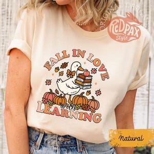 May include: A natural-colored t-shirt featuring a graphic with the text "FALL IN LOVE with LEARNING." The design includes a white duck holding books and an apple, surrounded by pumpkins and fall leaves. The Red Pax Style logo is visible.