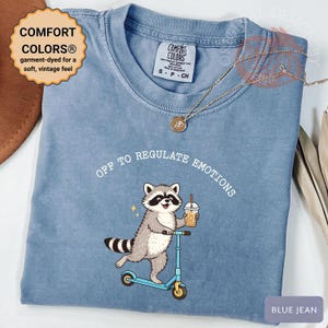May include: Blue jean colored t-shirt with a raccoon graphic riding a scooter and holding a drink. The text "OFF TO REGULATE EMOTIONS" is above the raccoon. The shirt has a vintage feel and is garment-dyed for softness.