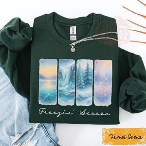 May include: Forest green sweatshirt with a winter landscape design. The design features four rectangular panels depicting a frozen lake, waterfall, snowy trees, and a field of snow-covered flowers. The words "Freezin' Season" are written in cursive below the panels.