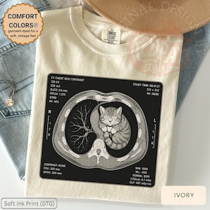 Könnte beinhalten: Elfenbeinfarbenes T-Shirt mit einer Grafik einer Katze in einem CT-Scan. Das Shirt trägt den Text "COMFORT COLORS®" und "Soft Ink Print (DTG)". Das Kleidungsstück ist für ein weiches, vintage-Feeling gefärbt.