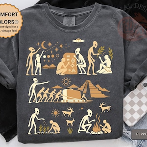May include: A charcoal gray t-shirt featuring a light tan graphic print of extraterrestrial beings interacting with ancient humans. The design depicts scenes of stargazing, stone carving, pyramid construction, farming, and fire-making in a stylized manner.