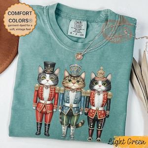 Op de afbeelding: Lichtgroen Comfort Colors T-shirt met een afbeelding van drie katten in speelgoedsoldatenuniformen. De katten dragen rode, blauwe en zwarte outfits met gouden details. Het shirt heeft de tekst "COMFORT COLORS" en "Light Green".