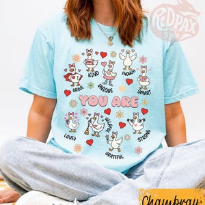 May include: Pale blue t-shirt featuring cartoon geese with various positive attributes. The words "YOU ARE" are above the geese, each accompanied by a different word such as "brave" and "kind." The shirt has short sleeves.