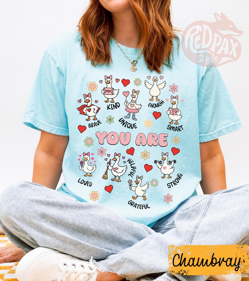 May include: Pale blue t-shirt featuring cartoon geese with various positive attributes. The words "YOU ARE" are above the geese, each accompanied by a different word such as "brave" and "kind." The shirt has short sleeves.