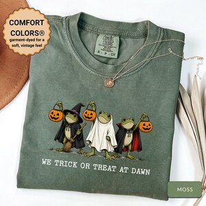 Puede incluir: Una camiseta de manga corta color verde musgo con un gráfico de tres ranas disfrazadas de Halloween sosteniendo cubos de calabaza. El texto dice WE TRICK OR TREAT AT DAWN. La camiseta tiene una etiqueta Comfort Colors en el cuello.