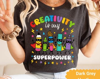 Könnte beinhalten: Dunkelgraues Heather-T-Shirt mit einer farbenfrohen Grafik. Die Grafik zeigt den Text "CREATIVITY is my SUPERPOWER" über Cartoon-Bleistiften, einem Pinsel, einem Lineal und einem Buntstift, alle mit Umhängen. Sterne und Blitze umgeben den Text.