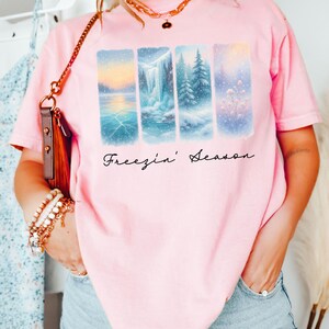 Puede incluir: Camiseta rosa claro con un dise&ntilde;o de paisaje invernal. El dise&ntilde;o presenta cuatro paneles rectangulares que representan escenas nevadas con el texto "Freezin' Season". La palabra "Blossom" est&aacute; en un rect&aacute;ngulo amarillo en la parte inferior.
