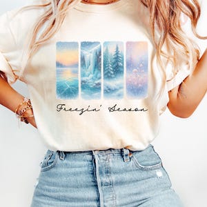 Puede incluir: Sudadera de color marfil con un dise&ntilde;o de paisaje invernal. Cuatro paneles verticales muestran escenas nevadas: un lago, una cascada y &aacute;rboles. Las palabras "Freezin' Season" est&aacute;n escritas en cursiva debajo.