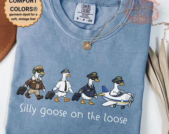 May include: A blue Comfort Colors t-shirt with a fun design. It shows four cartoon geese in pilot uniforms, each with a suitcase, and the text "Silly goose on the loose". A gold necklace is draped across the shirt. The shirt is garment-dyed.