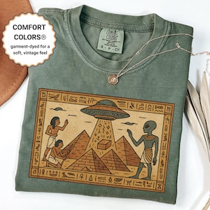 May include: Green Comfort Colors t-shirt featuring an Egyptian-themed graphic with pyramids, hieroglyphs, and a UFO. The shirt has the text "COMFORT COLORS® garment-dyed for a soft, vintage feel". A silver necklace with a pendant is draped across the shirt.