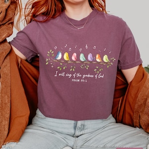 May include: A berry-colored short-sleeve t-shirt featuring a graphic of eight colorful birds perched on a leafy branch with musical notes above them. The text reads, "I will sing of the goodness of God" followed by "PSALM 89:1" in white lettering.