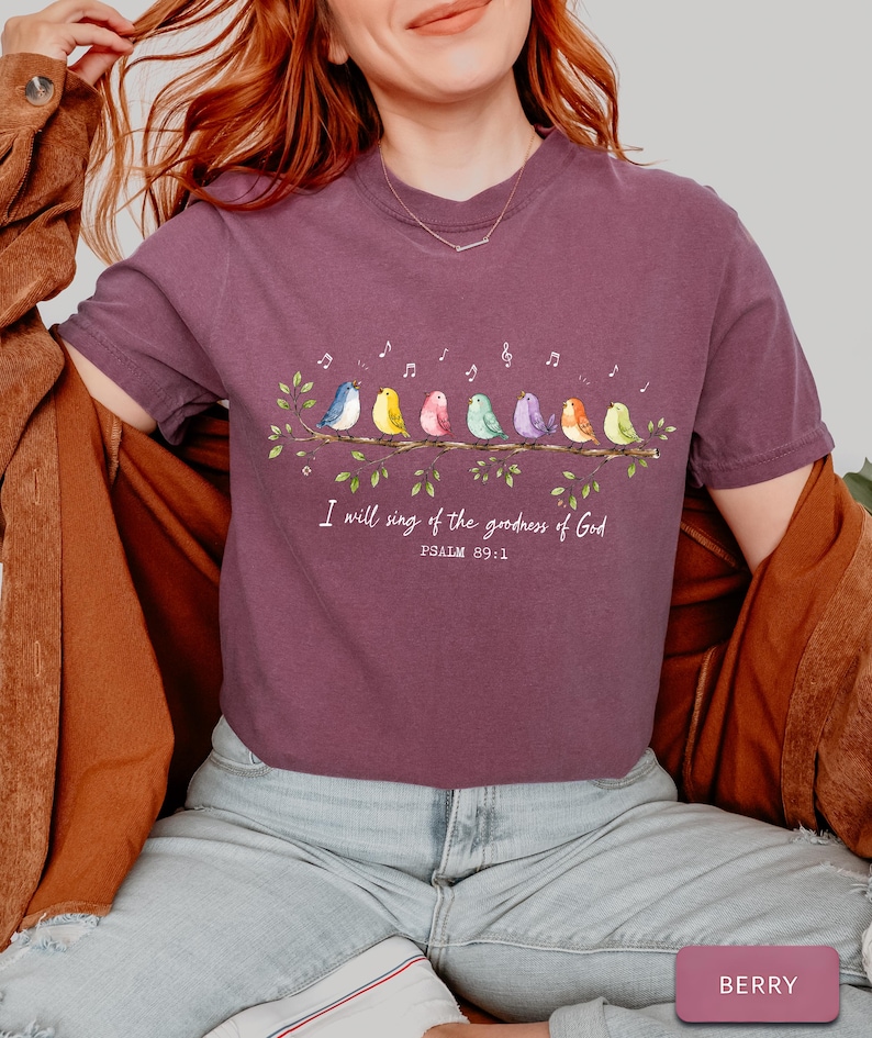 May include: A berry-colored short-sleeve t-shirt featuring a graphic of eight colorful birds perched on a leafy branch with musical notes above them. The text reads, "I will sing of the goodness of God" followed by "PSALM 89:1" in white lettering.