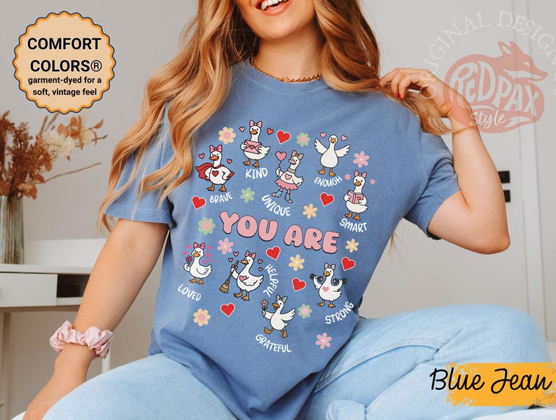 May include: Blue jean coloured t-shirt featuring cartoon geese and the words "YOU ARE" surrounded by positive affirmations. The garment-dyed shirt has a soft, vintage feel.