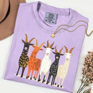 May include: A lilac Comfort Colors t-shirt featuring a graphic of five colorful goats. The goats are black, orange, white, and cream, with detailed horns. A gold star pendant necklace and a brown hat are also visible.